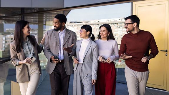 A group of five people are walking together in an office building. They are engaged in conversation and have happy expressions on their faces.