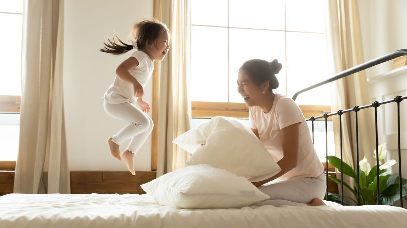 Mom smiling as her daughter has fun jumping on the bed