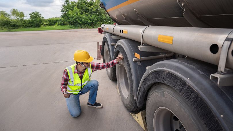 A man outside in a safety vest squats down to inspect the wheel nuts of a commercial transport truck trailer