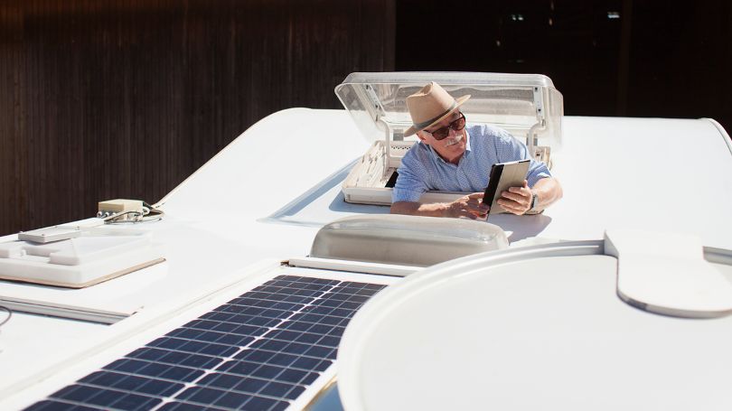 A senior man testing the solar panels atop his camper van with a tablet. 