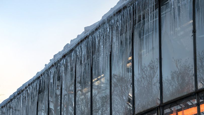 Accumulation of snow on the roof of commercial building with big icicles hanging outside glass walls.