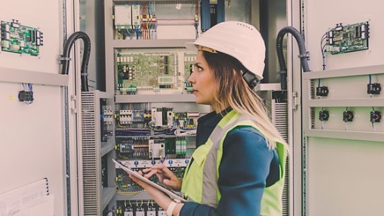 Woman engineer wearing white helmet, checking a control panel and writing notes with digital tablet in the control room.