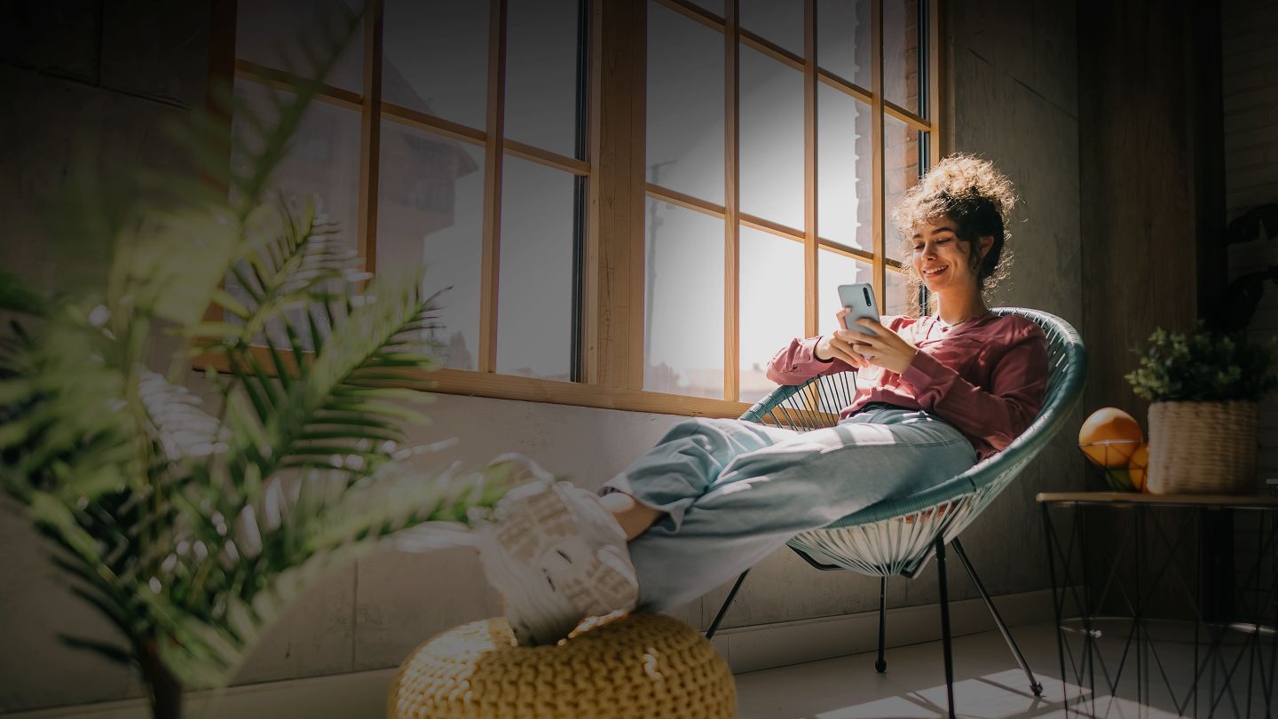 Une femme assise confortablement sur une chaise près de la fenêtre utilisant un téléphone intelligent.