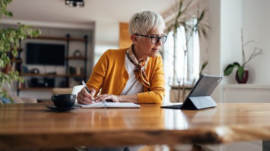 A mature woman sitting at a desk writing on a notepad while looking at her tablet.