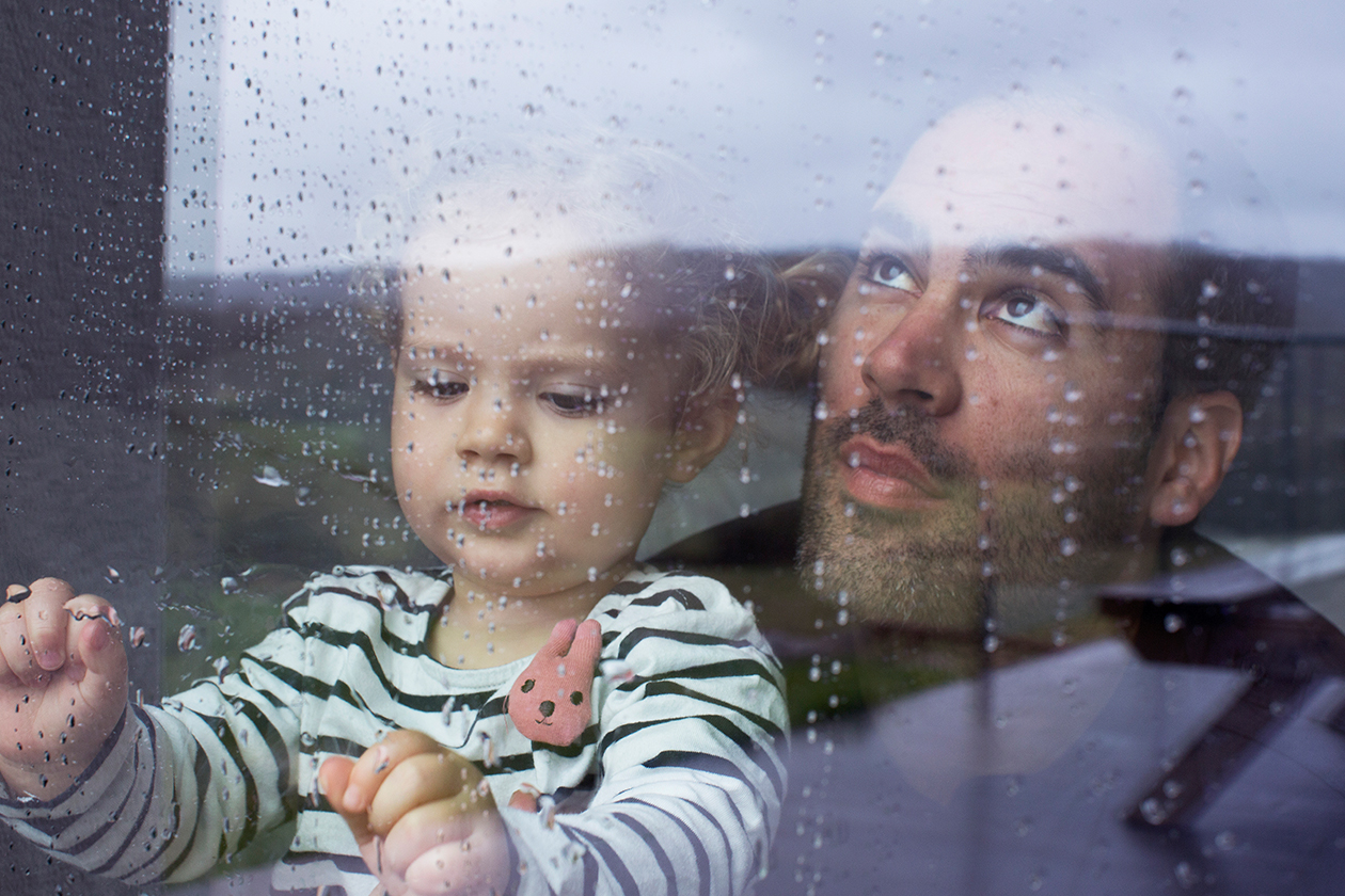 Man with a young child looking through a window watching the rain